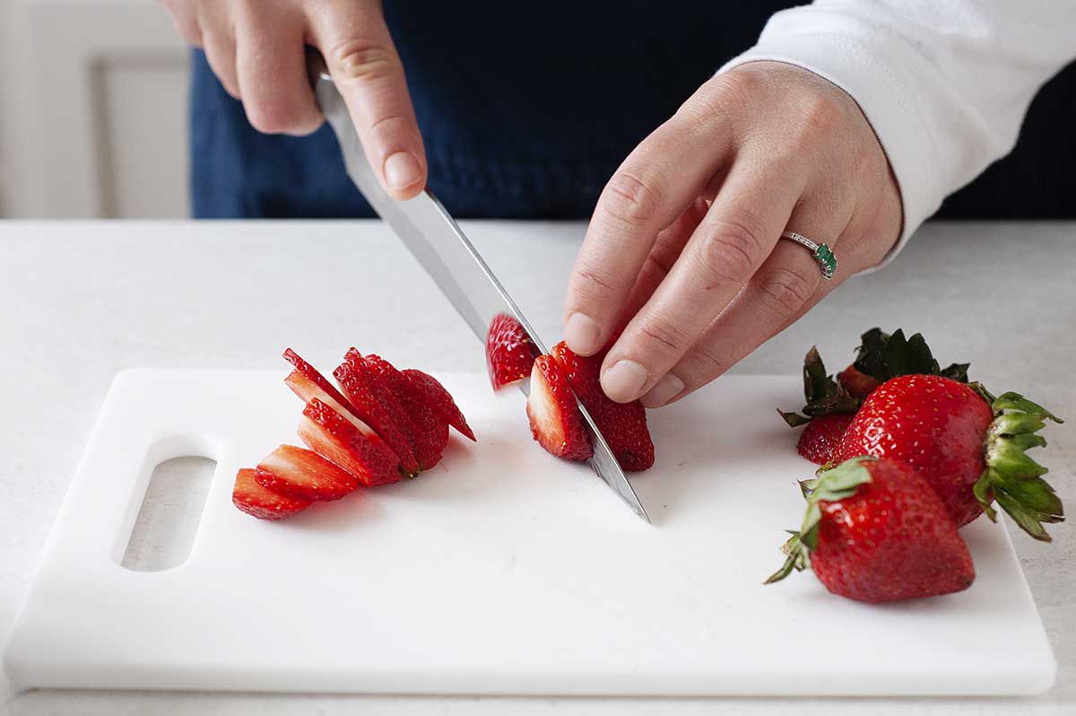 Strawberries being sliced on a white cutting board.