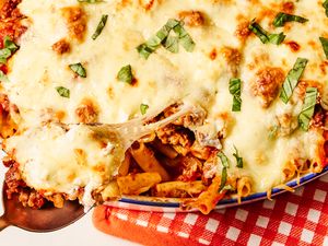 A closeup of baked ziti topped with melted cheese and herbs in a dish with a serving spoon lifting a portion from the dish