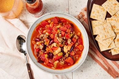 Bowl of copycat Wendy's chili at a table setting with a bowl of saltines on a brown and white kitchen towel, a bottle of tabasco, a drink, and a white table napkin