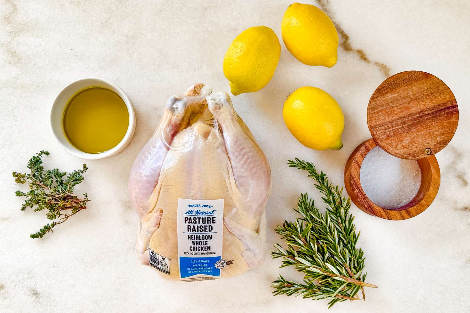 A raw chicken still in its packaging, three lemons, a wooden salt cellar, sprigs of fresh rosemary and thyme, and a white ramekin of olive oil all on a marble counter