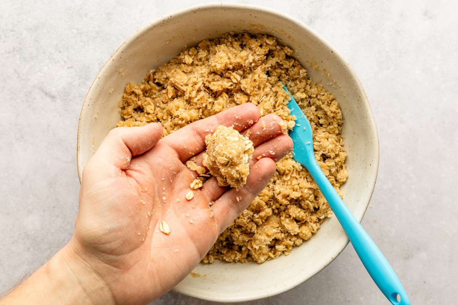 A small ball of dough for the Anzac Biscuits recipe shaped in a hand above a mixing bow with the rest of the dough