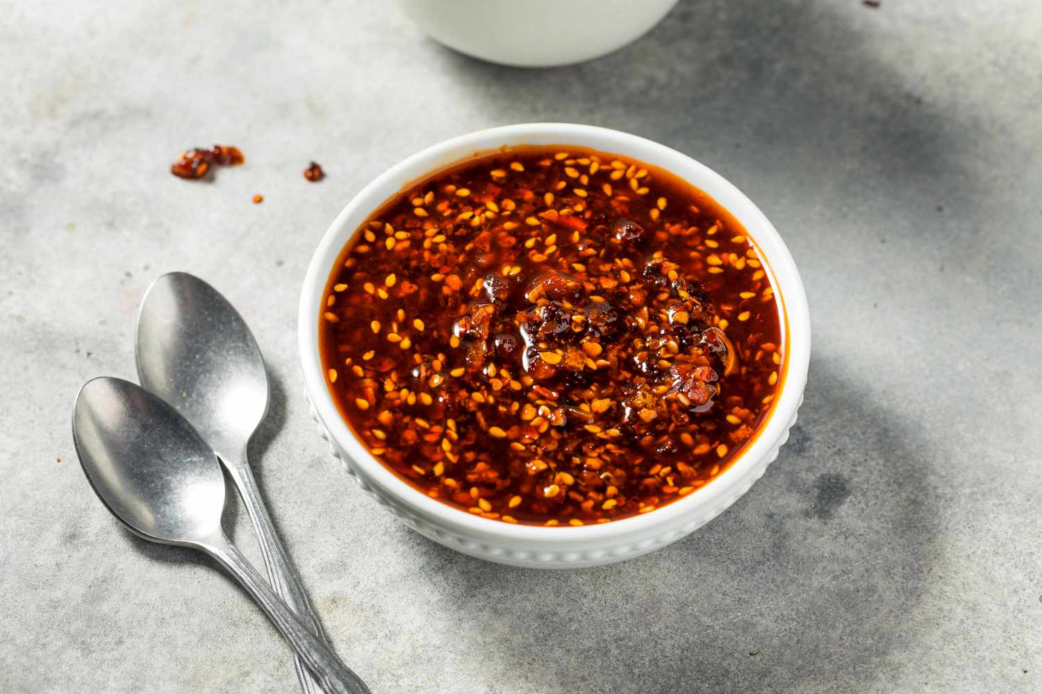 A bowl of chili crisp beside two spoons on a gray surface