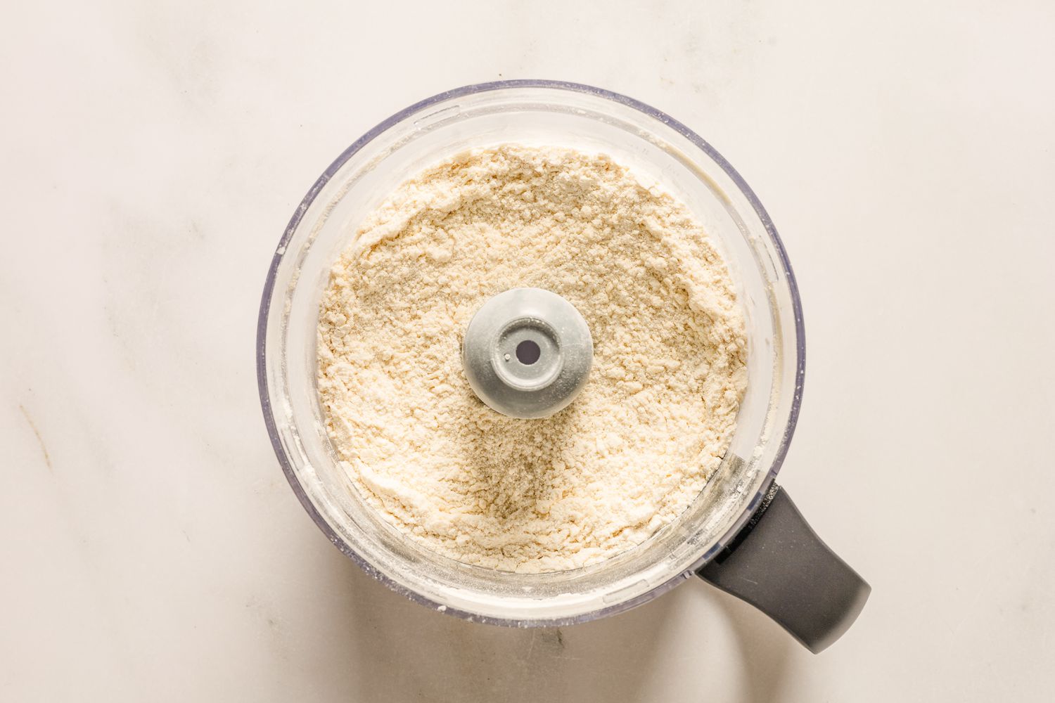 An overhead view of a food processor bowl with the butter and dry ingredients after processing to resemble coarse sand for Sand Tarts recipe