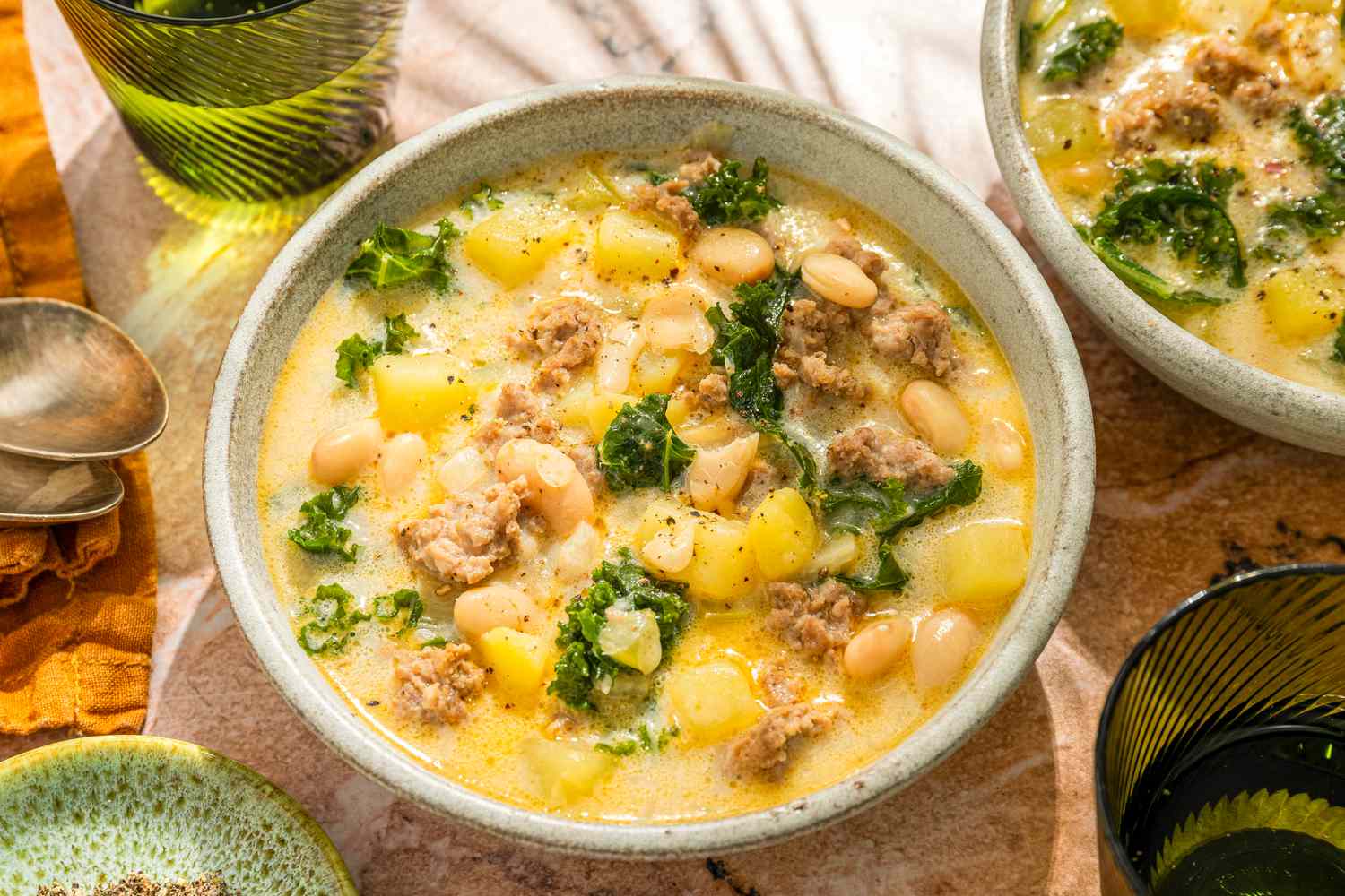 Side angle view of two bowls with a cream based soup with beans, sausage, kale and potatoes, and two green water glasses