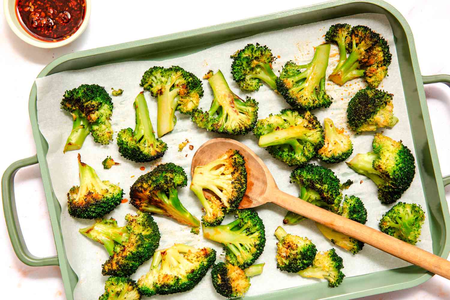 Overhead view of a baking sheet of chili crisp coated broccoli florets on parchment paper with a serving spoon next to a small bowl of chili crisp