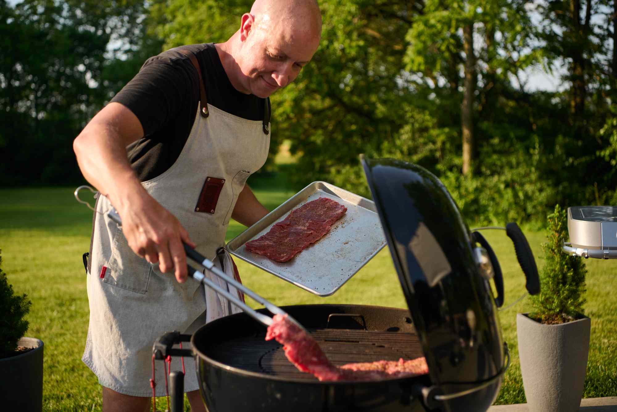 A person grilling meat outdoors using a round charcoal grill, holding tongs and a tray with slices of meat