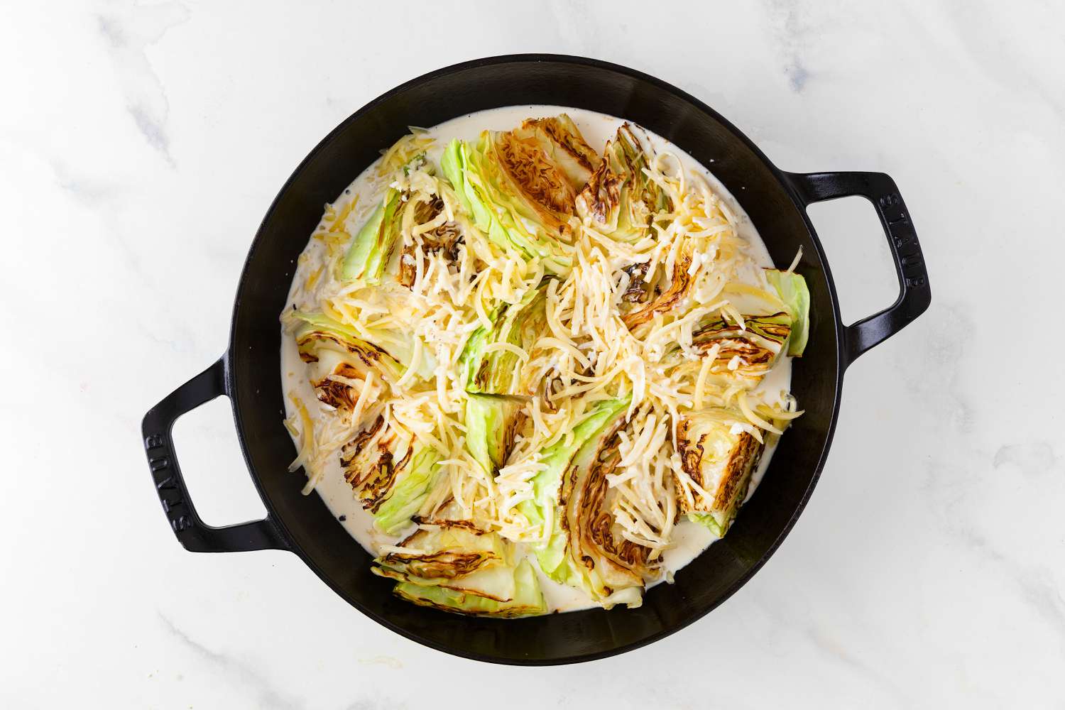 Overhead view of browned cabbage in a cast iron skillet after adding the cream, garlic, salt, black pepper, and shredded gruyere for Cabbage Au Gratin recipe