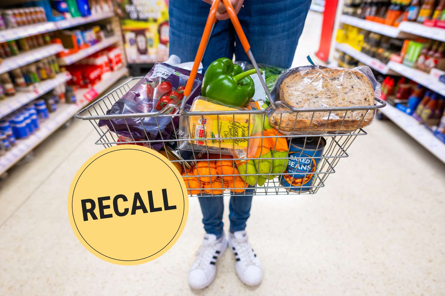 A person from the waist down wearing jeans and Adidas sneakers standing in the middle of a grocery store aisle holding a full shopping basket. A "Recall" badge is superimposed over the photo.