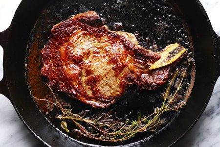 A cast iron skillet with steak cooked on the stove. Oregano sprigs are at the bottom of the pan. Oil and juices are visible in the pan.