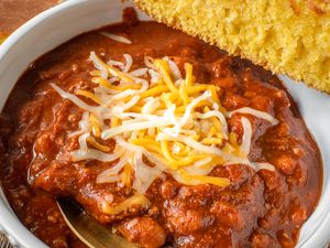 Closeup view of a white bowl of chili with cheese topping and spoon along with a slice of cornbread balanced on the edge of the bowl