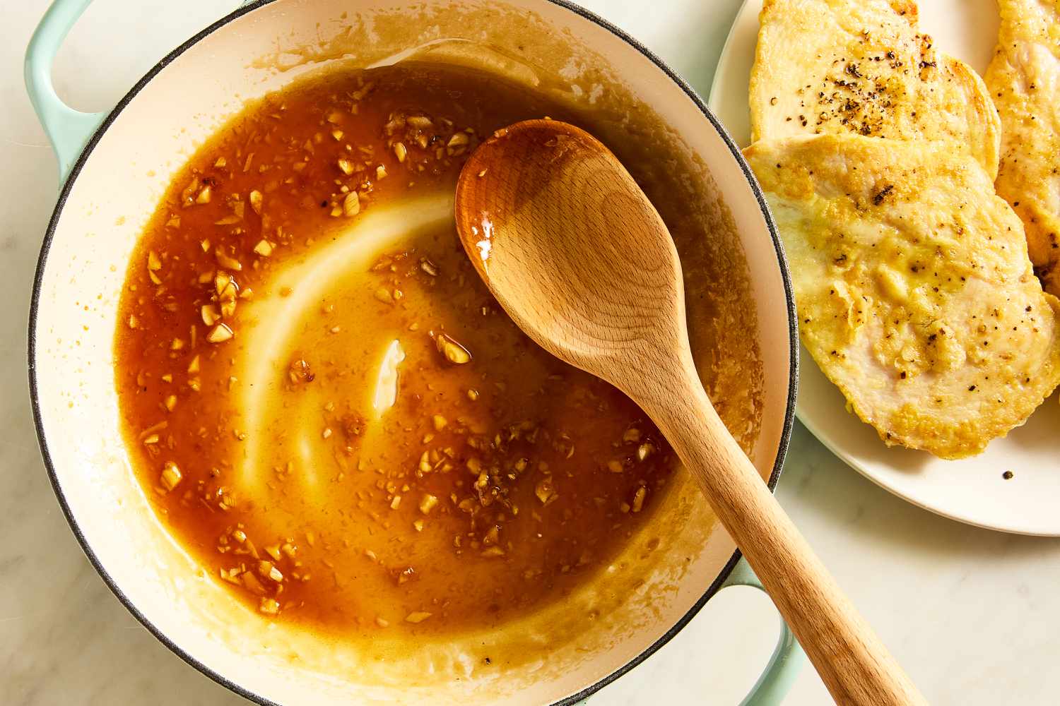 Overhead view of a teal dutch oven with a spoon and the soy sauce mixture next to a white plate of cooked chicken breasts for Honey Soy Chicken recipe
