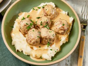 Bowl of copycat Ikea Swedish meatballs served over mashed potatoes, and in the surroundings, a table setting with a stack of plates and a sage table napkin