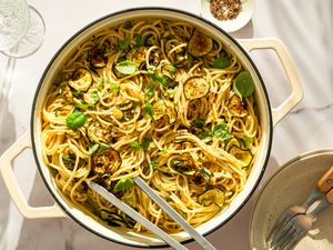 A pot containing spaghetti with zucchini slices and garnished with basil, alongside a plate, utensils, and condiments