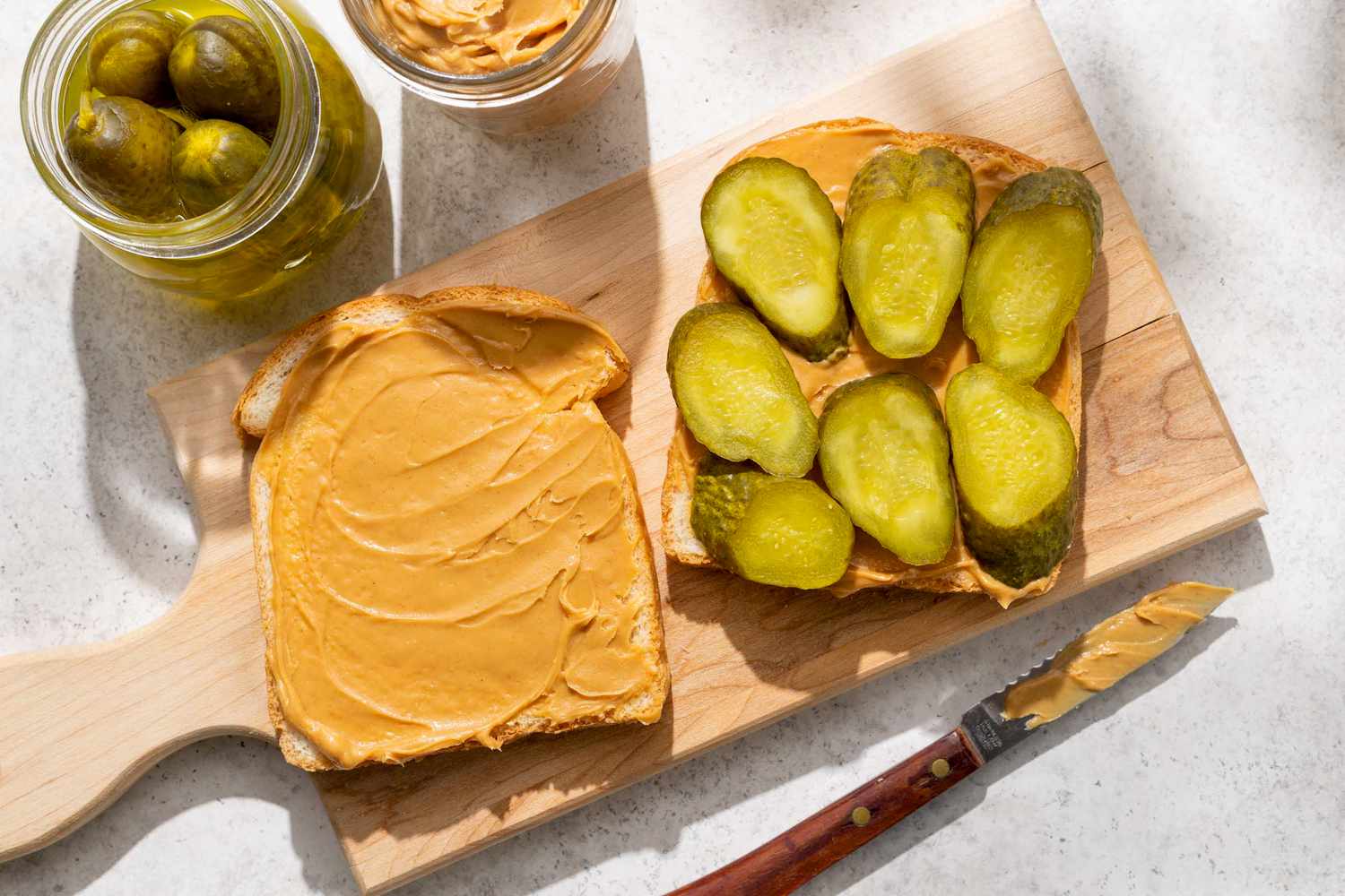 Peanut butter spread on bread with pickles on another slice knife and jar of pickles beside