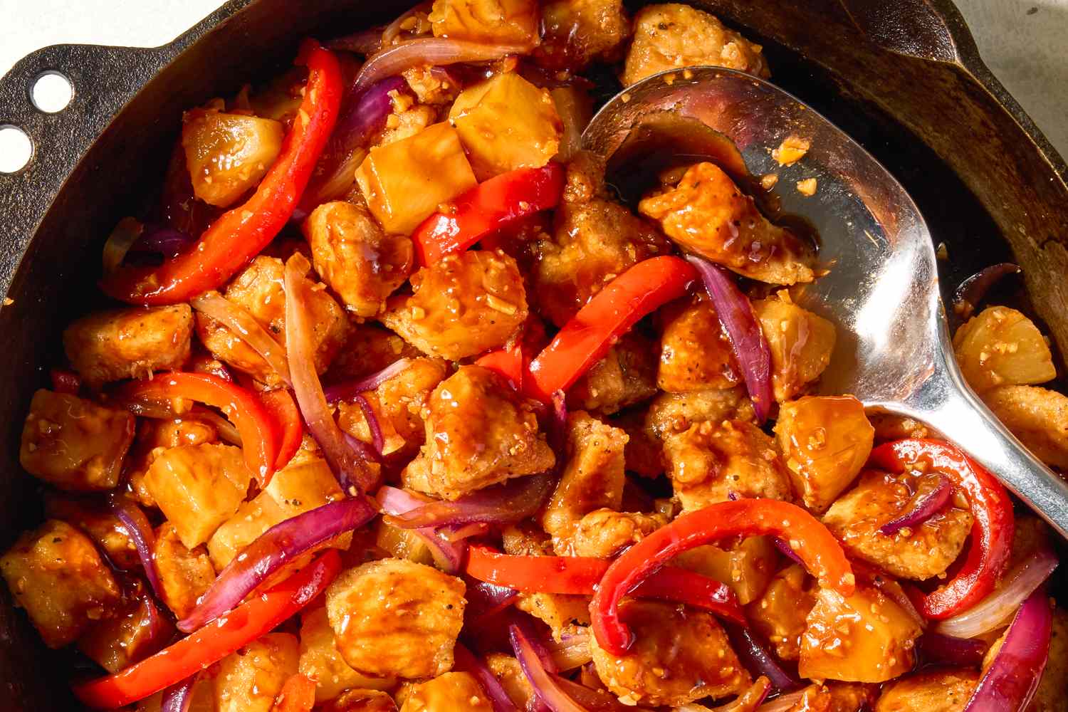 A skillet containing cooked chunks of chicken red bell peppers pineapple and red onions in a savory sauce