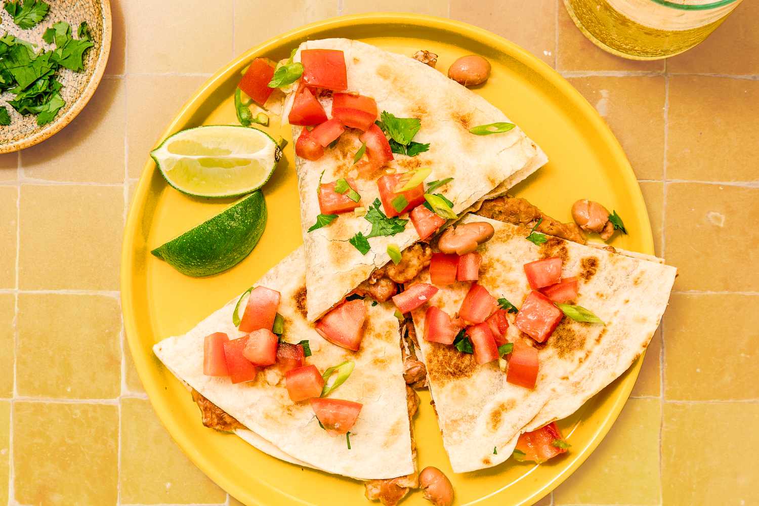 Three slices of a quesadilla on a plate along with lime slices at a table setting with a small plate of cilantro and a drinking glass
