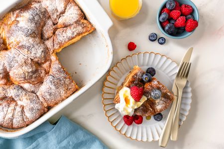 Overhead view of a white baking dish of Croissant French Toast casserole with a slice removed and placed on a small plate with a knife and fork along with a glass of orange juice and bowl of fruit