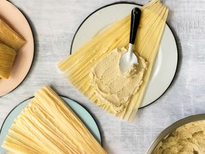 Bowl of Masa (Tamales Dough) Next to a Plate with a Corn Husk Smeared with Masa Using the Back of the Spoon and a Plate with More Corn Husks