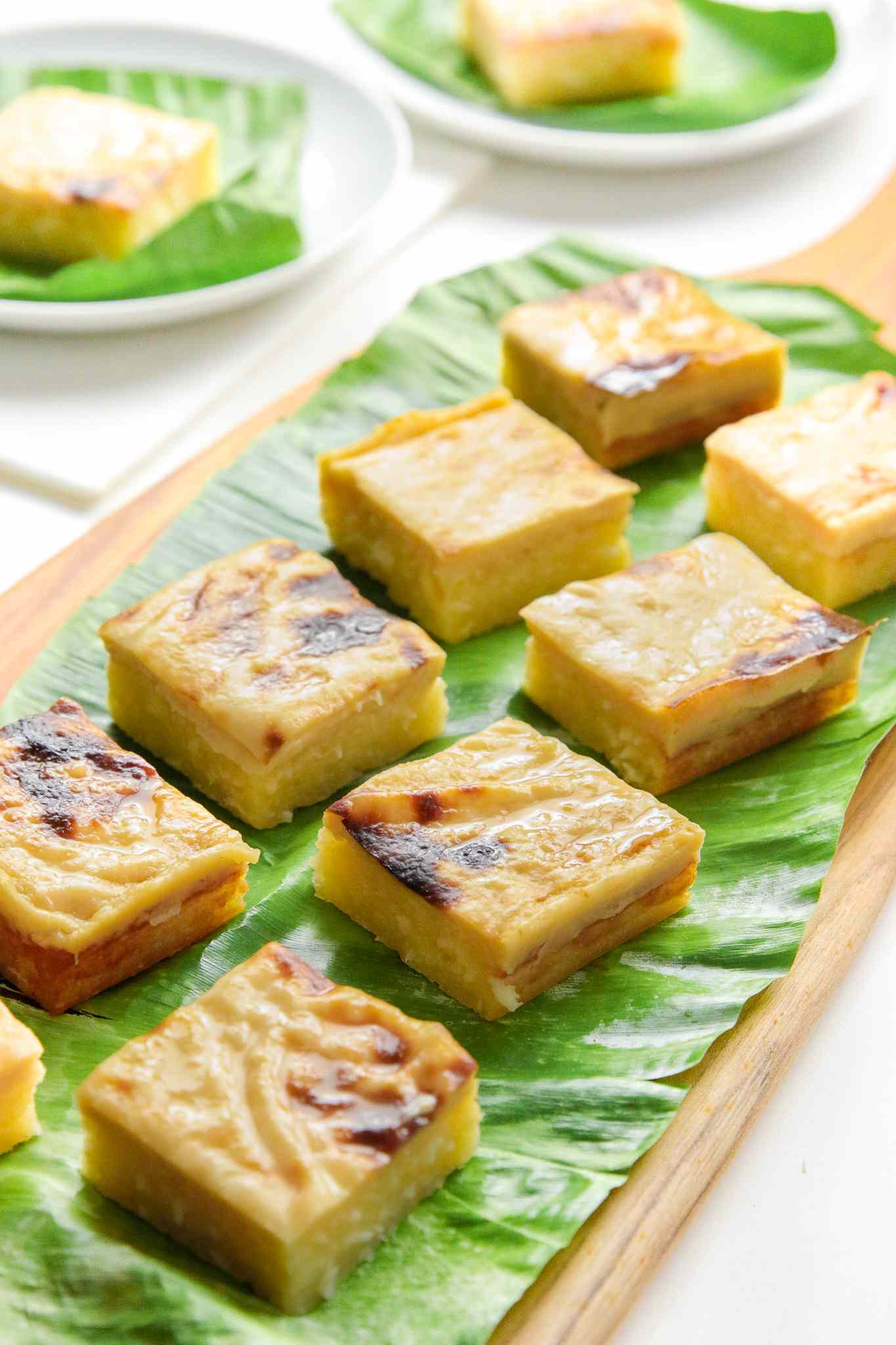 Cassava Cake Slices on a Banana Leaf Covered Wooden Tray, and in the Background, Individual Slices of Cake on Two Banana Leaf Covered Plates