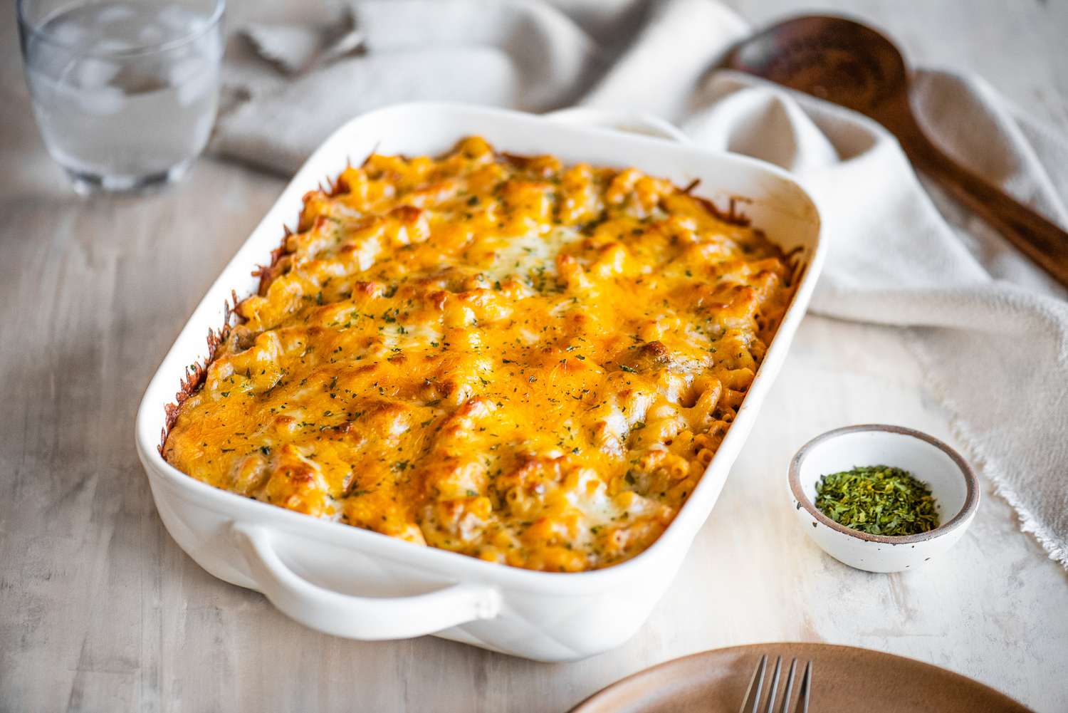 Cheeseburger Casserole in a Casserole Dish at a Table Setting (a Stack of Plates with Two Forks, a Spoon on the Counter, Bowl of Dry Parsley, a Table Napkin, and a Glass of Ice Water)