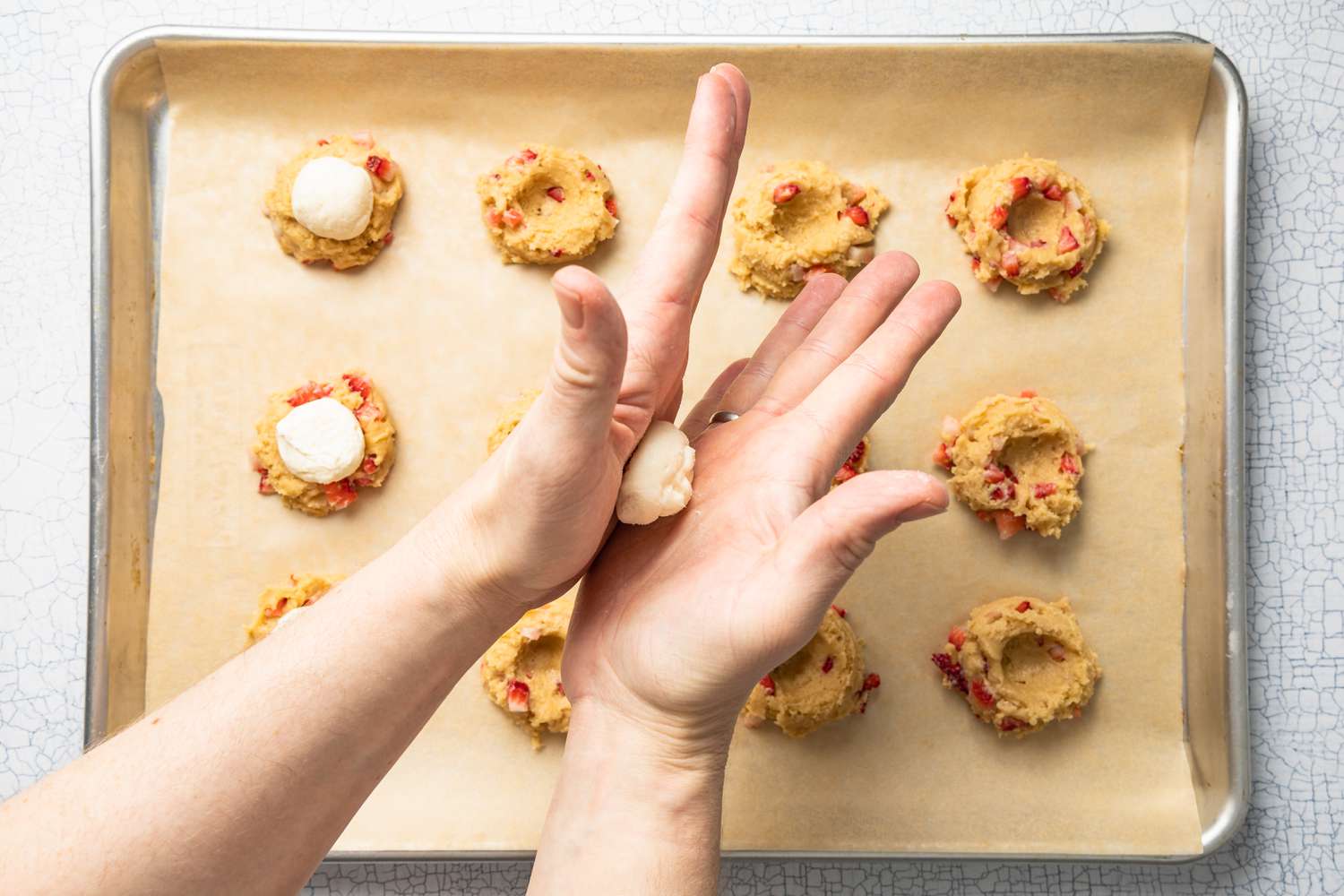 A Pair of Hands Rolling Frozen Cheesecake Filling Into a Neat Ball, and in the Background Below, a Parchment Paper Lined Baking Sheet With Dollops of Indented Cookie (Some With Cheesecake Filling Sitting in the Thumbprint)