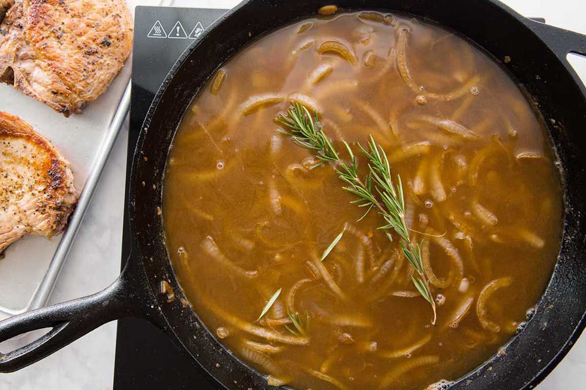 Onions with rosemary in a cast iron skillet filled with gravy and being stirred with a wooden spoon.