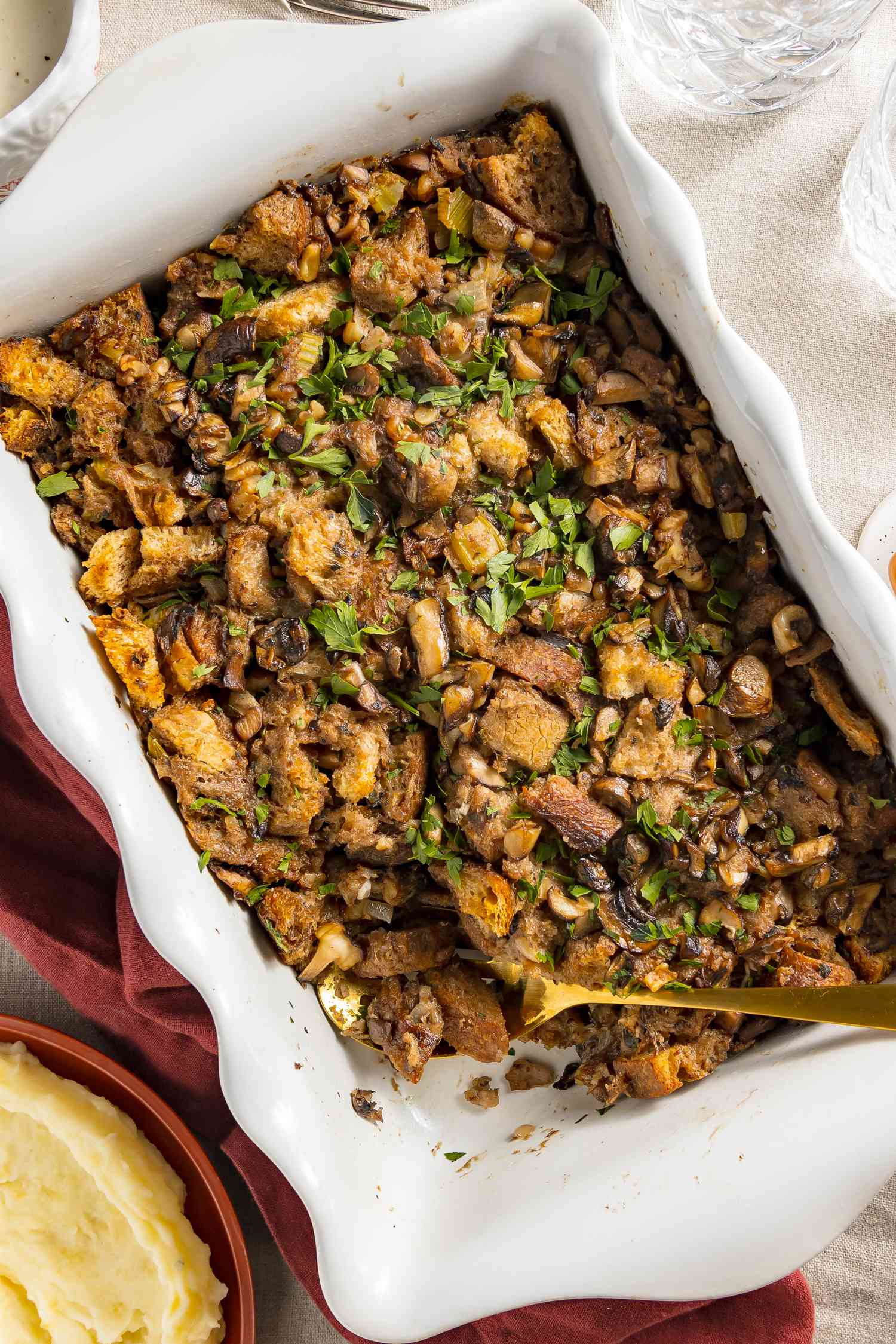 Table Setting: Vegan Mushroom Stuffing in a Casserole Dish Next to a Bowl of Mashed Potatoes, Two Glasses of Water, and a Table Napkin