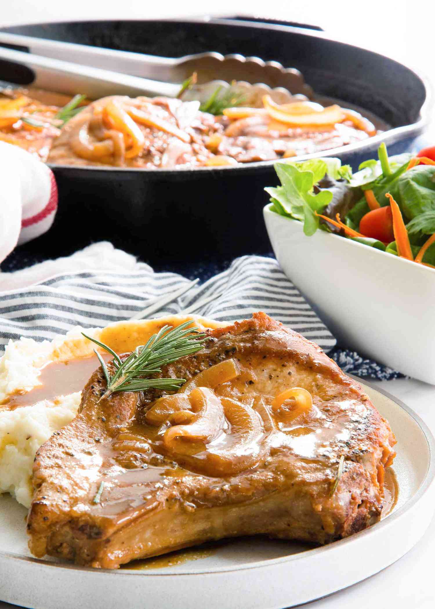 A gluten free smothered pork chop with a pile of mashed potatoes covered in gravy on an ivory plate with a striped linen nearyby and a side salad.
