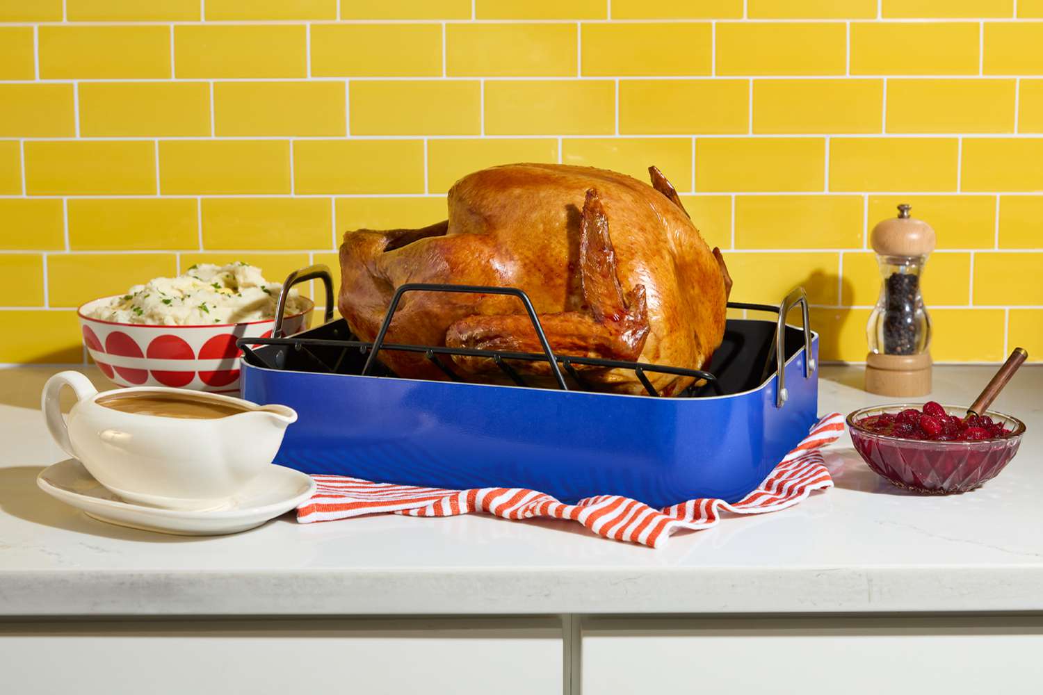 A golden brown turkey in a roasting pan on a kitchen counter surrounded by mashed potatoes, cranberry sauce, and gravy