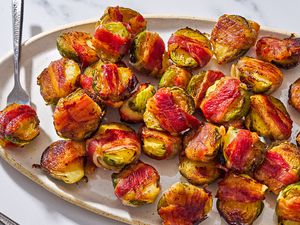 Closeup of bacon wrapped Brussels sprouts on a large oval serving plate, with a fork stuck into one of the sprouts