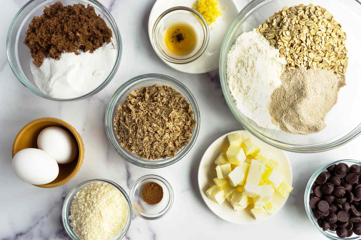 Ingredients for making cookies including eggs oats butter and sugar arranged in bowls on a countertop