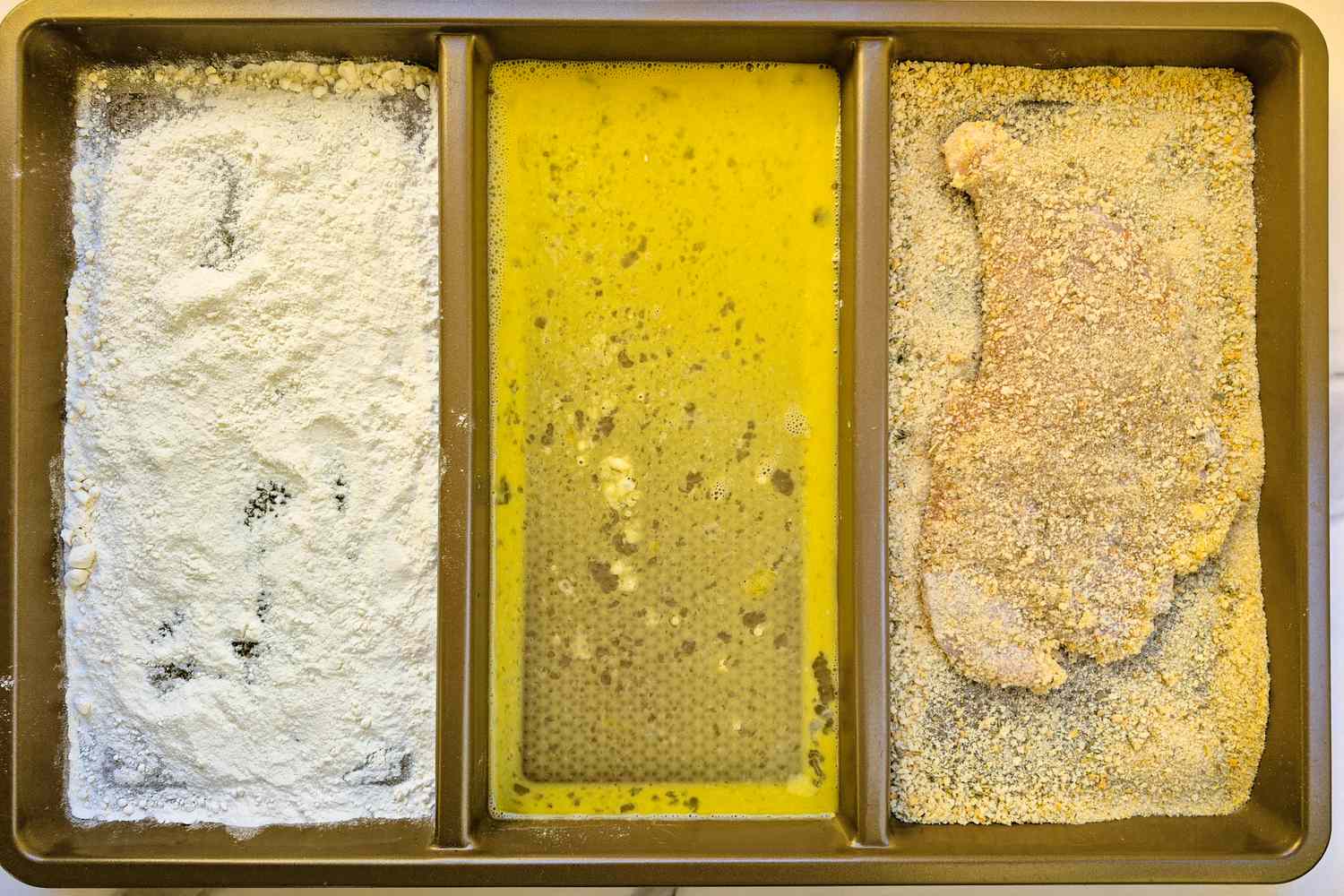 A breading station with sections featuring flour beaten egg and breadcrumbs used for preparing breaded chicken