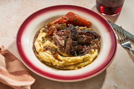A serving bowl of pot roast over mashed potatoes with carrots on the side