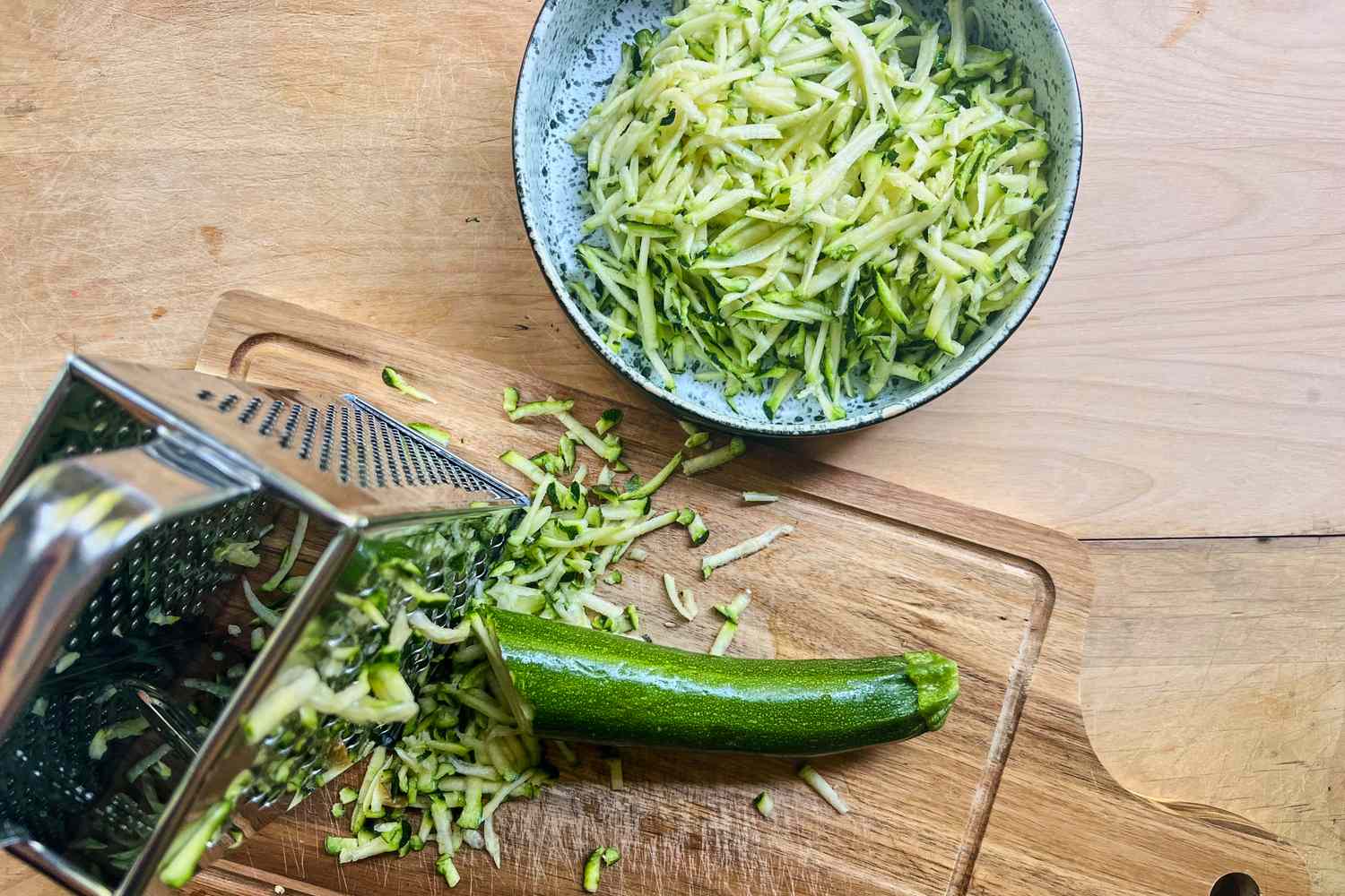 Grated zucchini spilling from a grater onto a cutting board with a bowl of shredded zucchini nearby