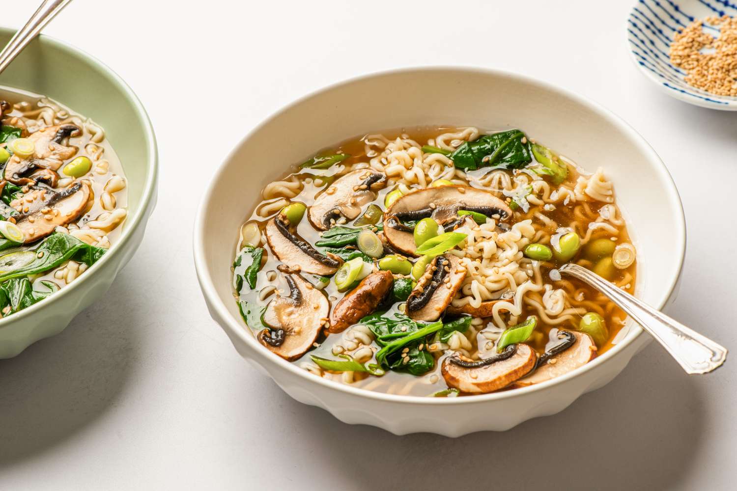 A bowl of mushroom noodle soup with greens and a spoon tofu and seasoning in surrounding bowls