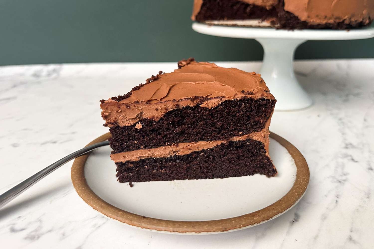 Slice of chocolate cake with frosting on a plate with the rest of the cake on a stand in the background