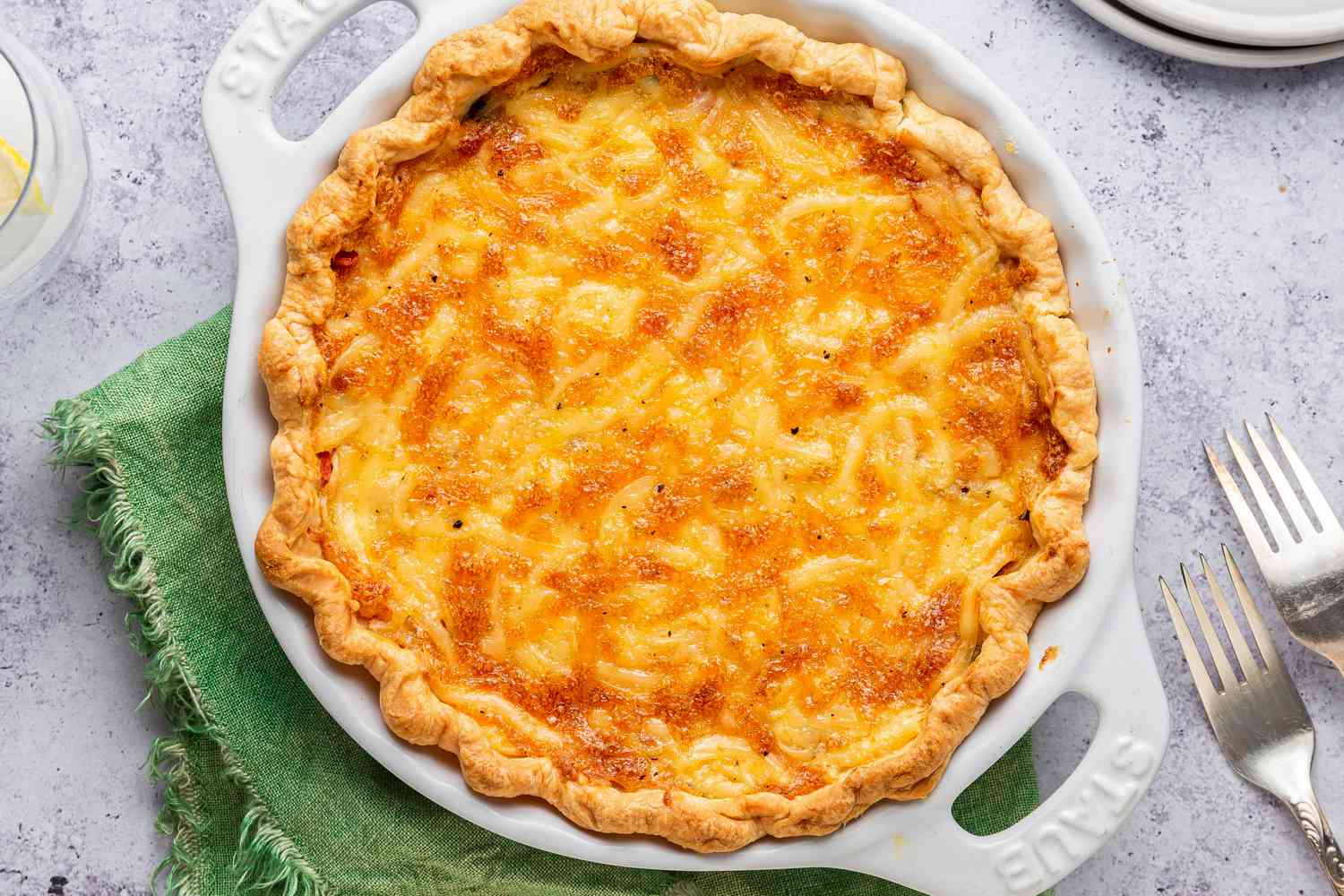 tomato pie in a pie pan at a table setting with utensils, a stack of plates, and a glass of water