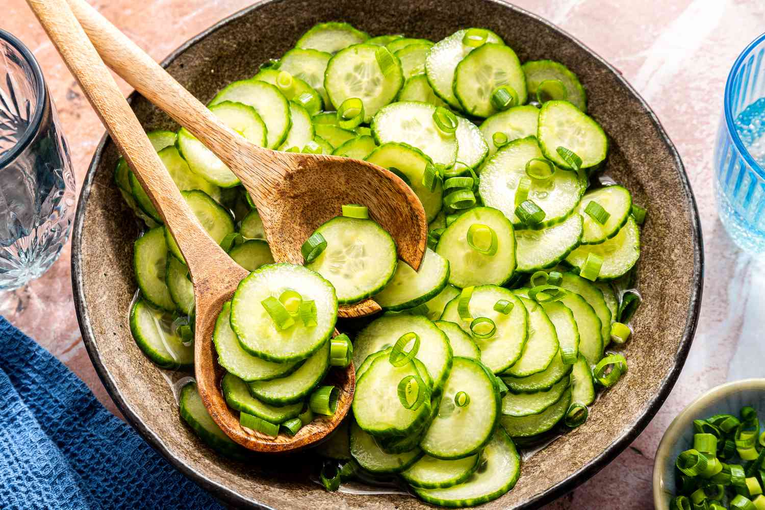 cucumber salad in a bowl with a pair of serving utensils at a table setting with a bowl of slcied scallions, a table napkin, and a glass of water 