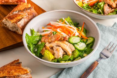 Banh mi salad and in the surroundings, another bowl, a wooden board with baguette slices, and a table napkin with a fork