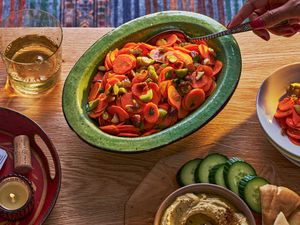A bowl of carrot salad on a coffee table, alongside hummus with cucumbers and pita and a glass of wine