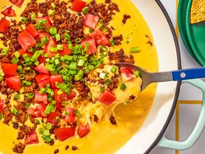 Queso dip with toppings of chopped tomatoes green onions and chorizo served with a spoon and tortilla chips on the side