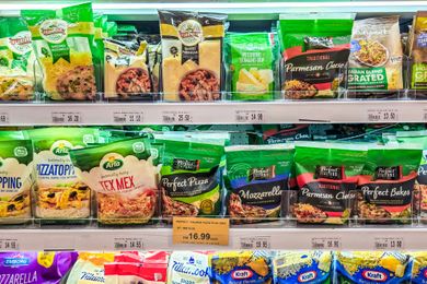 A grocery store shelf displaying various brands and types of shredded cheese in green and colorful packaging