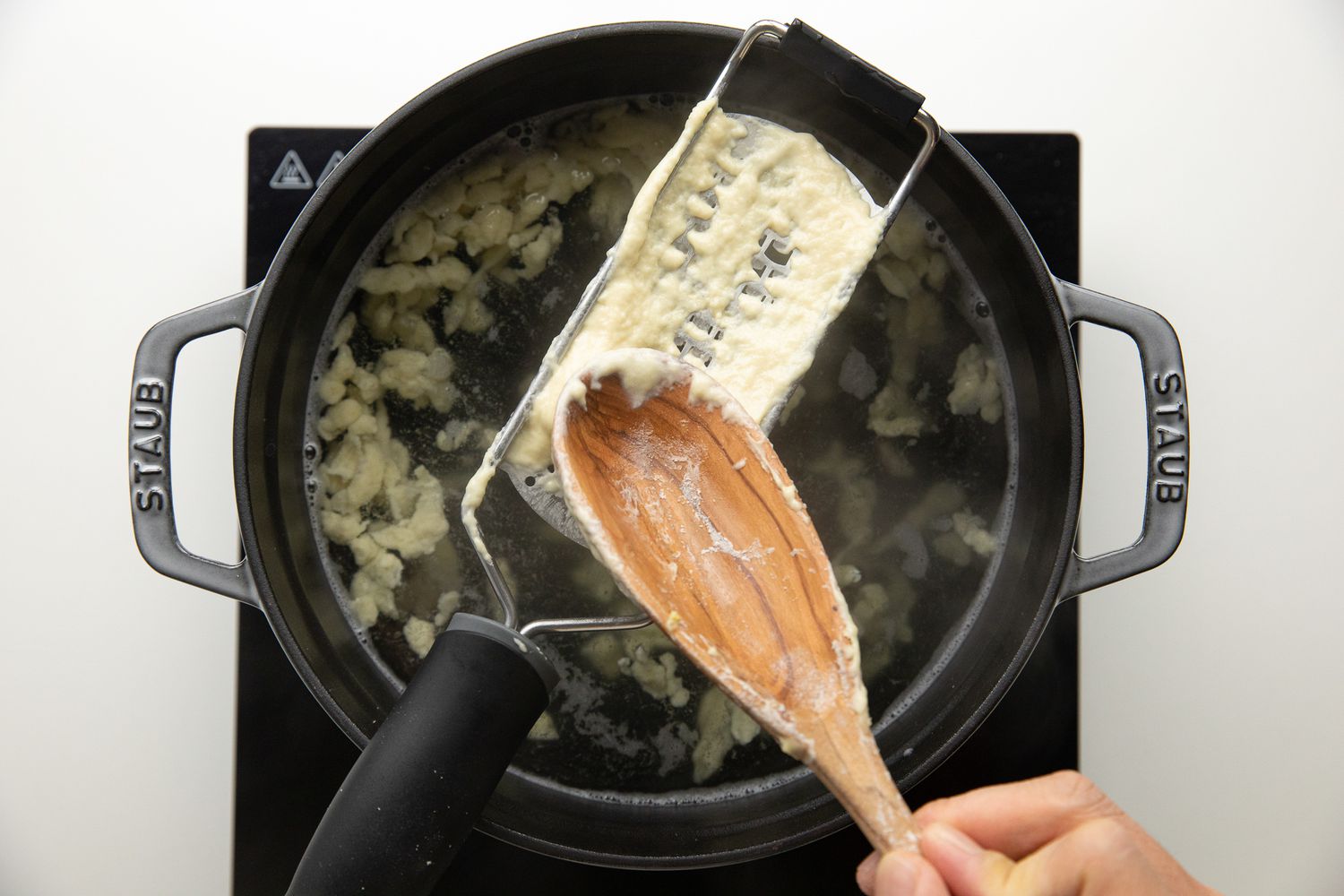 Wooden Spoon Pressing the Homemade Spaetzle Dough Through the Cheese Grater into the Dutch Oven Filled with Water 