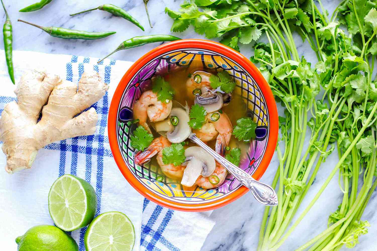 Thai soup with shrimp in a brightly patterned bowl with a silver spoon inside. Sliced limes, ginger, peppers and cilantro stems suround the bowl of light broth, shrimp, mushrooms and cilantro. The bowl is on a marble counter with a blue striped linen to the left of the bowl.