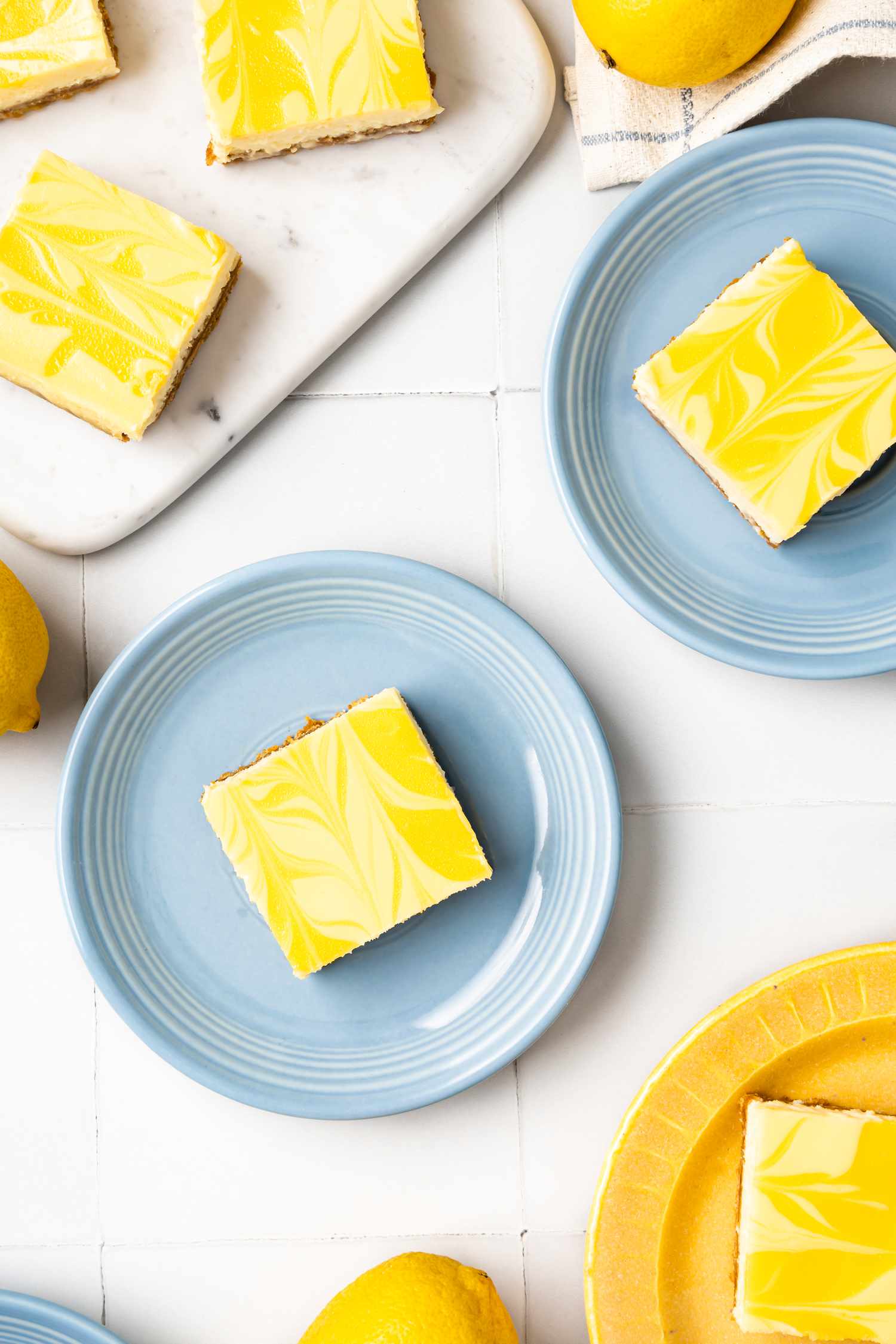 Lemon Cheesecake Bars on Three Plates and More on the Marble Board Next to the Plates, and on the Counter, Lemons and a Kitchen Towel