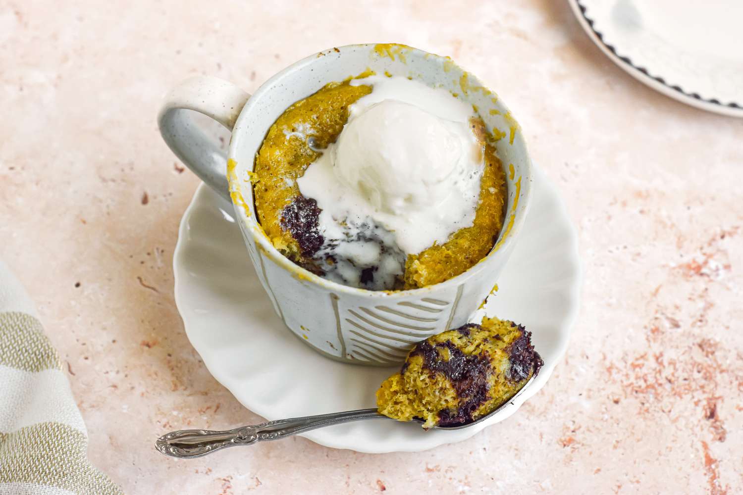 Mug of Banana Bread Chocolate Chunk Mug Cake Topped with a Dollop of Ice Cream and Spoonful of Cake Resting on a Saucer (Mug on the Saucer Next to the Spoon)