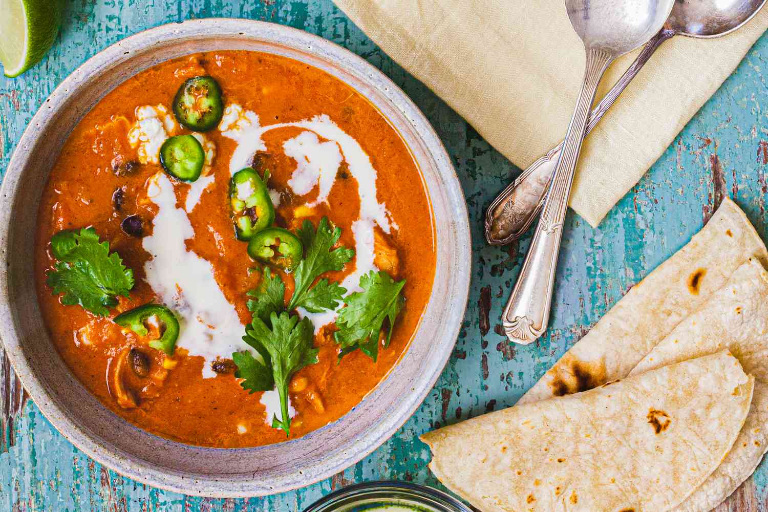 Creamy Chicken Enchilada Soup Topped with Jalapenos, Cilantro, and Crema in a Bowl at a Table Setting with Two Spoons on a Table Napkin and Tortillas