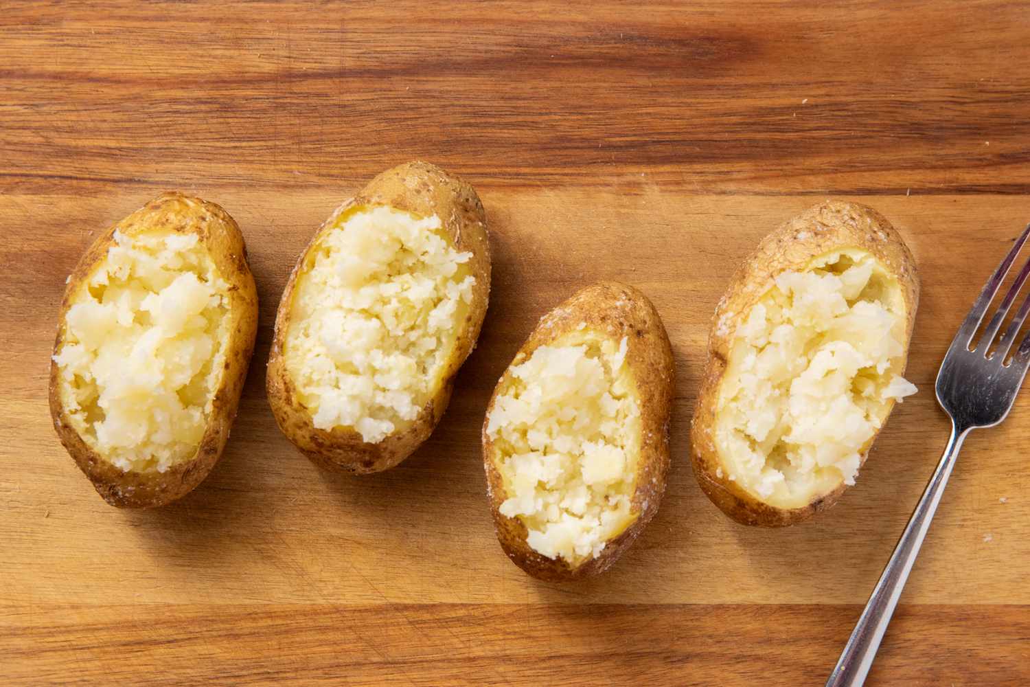 Four baked potato halves with the insides gently fluffed placed side by side on a wooden surface accompanied by a fork for Loaded Chopped Cheese Baked Potatoes
