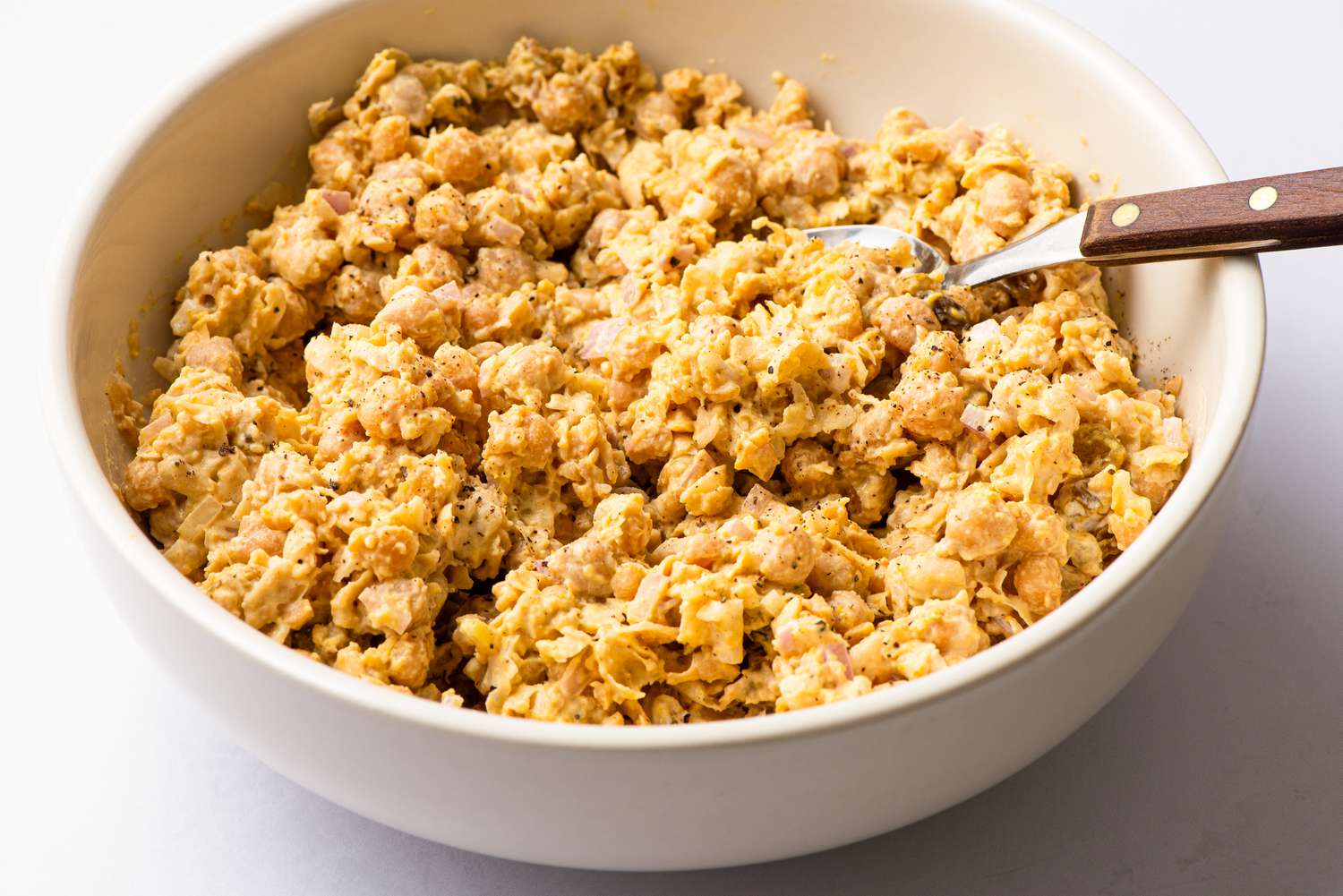 Angled view of a white bowl of curried chickpea salad with a spoon all on a white background