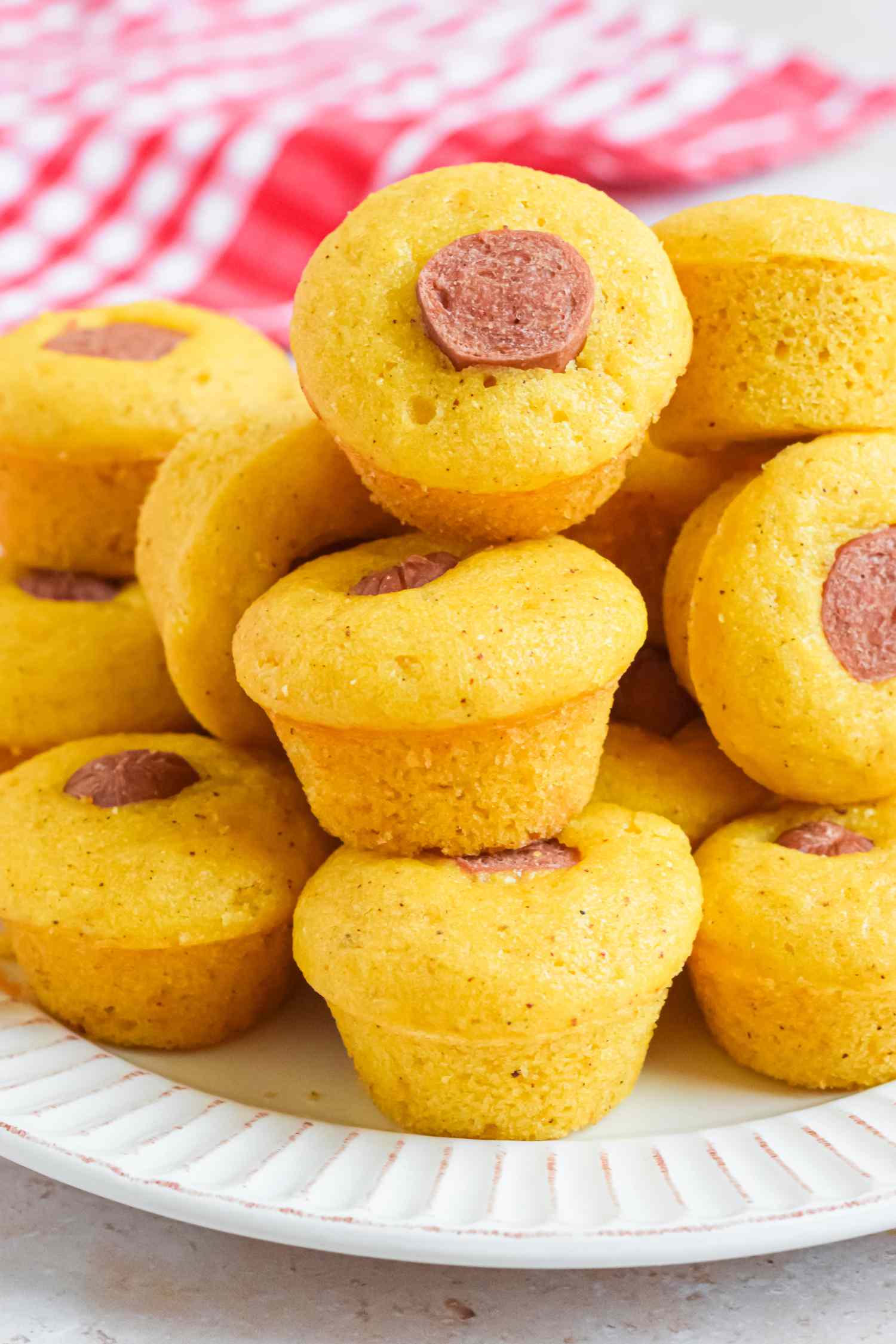 Pile of Corn Dog Muffins Stacked on a Plate, and in the Background, a Red and White Checkered Kitchen Towel 